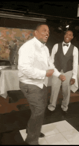 a man in a tuxedo is dancing on a dance floor with two waiters behind him .