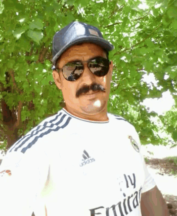 a man wearing sunglasses and a fly emirates shirt stands in front of a tree