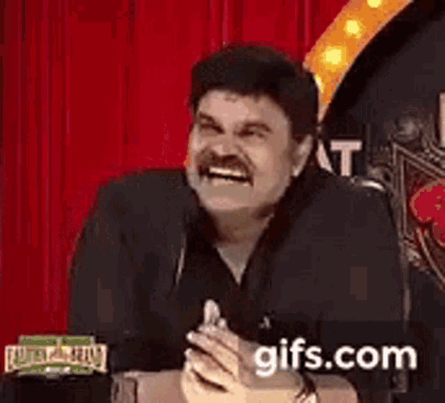 a man with a mustache is laughing into a microphone while sitting in front of a red curtain .