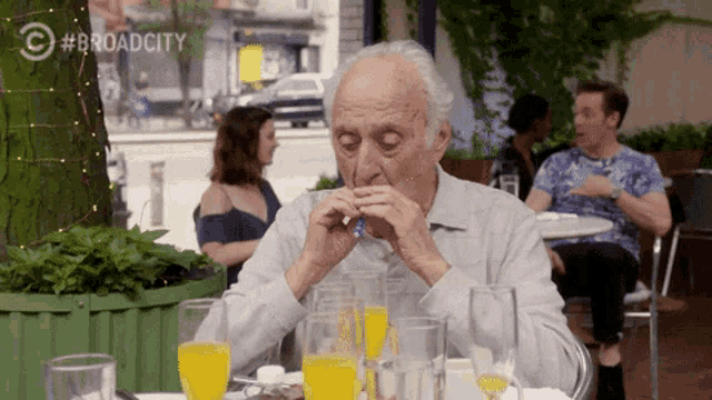 an older man sits at a table with glasses of orange juice in front of a sign that says broadcity