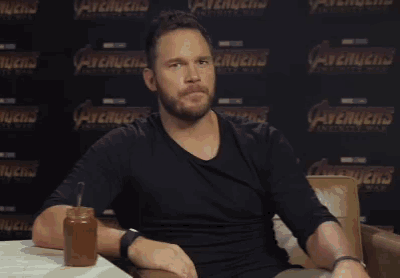 a man with a beard is sitting in front of a wall with the word avengers on it .