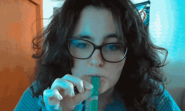 a woman wearing glasses and a blue sweater is eating a green stick