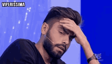 a man with a beard holds his hand to his forehead in front of a microphone with the word viperissima above him