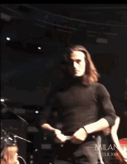a man with long hair wearing a black turtleneck sweater is standing on a stage in milano