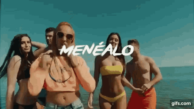 a group of people in bikinis are dancing on a beach with the word menealo written above them .