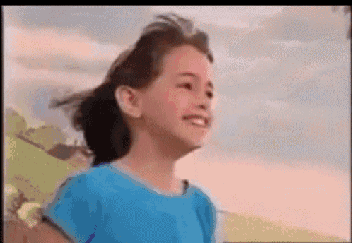 a little girl in a blue shirt is smiling and looking up at the sky .