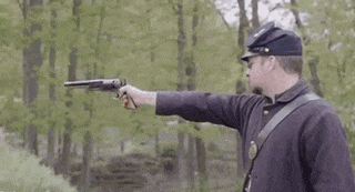 a man in a military uniform is holding a gun and pointing it at something in the woods .