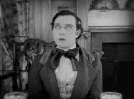 a man in a suit and bow tie is sitting in front of a fireplace in a black and white photo .