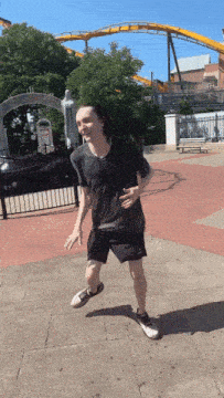 a man in a black shirt and black shorts is dancing in front of a roller coaster