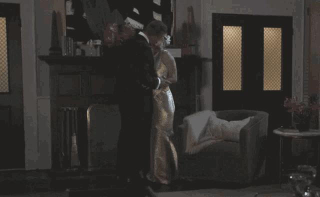 a man in a tuxedo and a woman in a gold dress are kissing in a living room