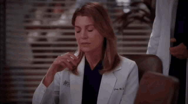 a woman in a grey 's anatomy lab coat sits in a chair