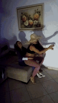 two women are wrestling on a couch in front of a framed painting of flowers
