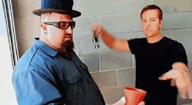 two men are standing next to each other in front of a brick wall . one man is wearing a top hat and sunglasses .