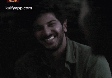 a man with a beard is smiling in a dark room and the website kulfyapp.com is visible in the corner