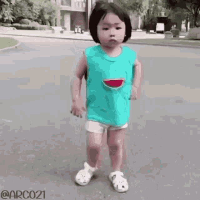 a little girl wearing a watermelon shirt and crocs is standing on a street .