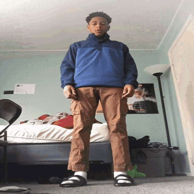 a young man wearing a blue jacket and brown pants is standing in front of a bed