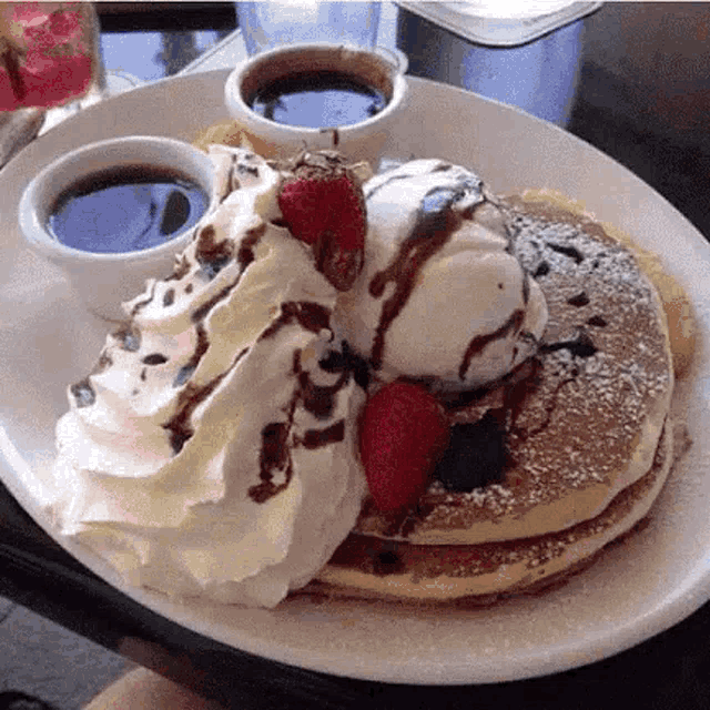 a person is holding a plate of pancakes with whipped cream , ice cream , strawberries and chocolate sauce .