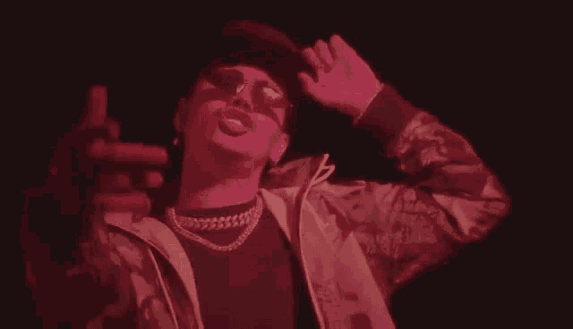a man wearing a hat , sunglasses , a necklace and a jacket is dancing in a dark room .