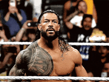 a shirtless wrestler with a tattoo on his chest stands in a rope ring