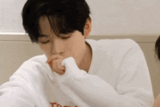 a young man in a white sweater is sitting at a table with his hand on his mouth .
