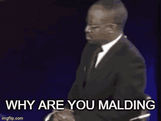 a man in a suit and tie is sitting down with the words `` why are you malding '' behind him .