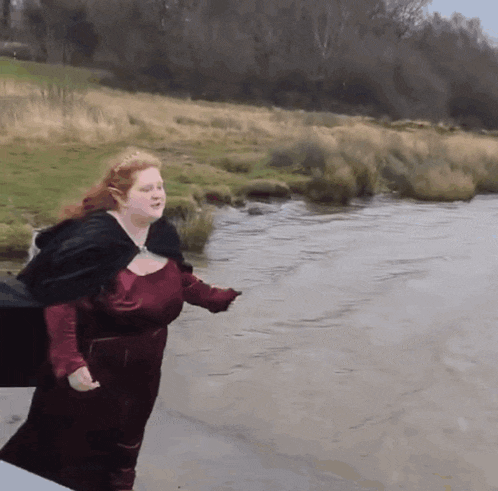 a woman in a red dress and black cape stands in front of a river