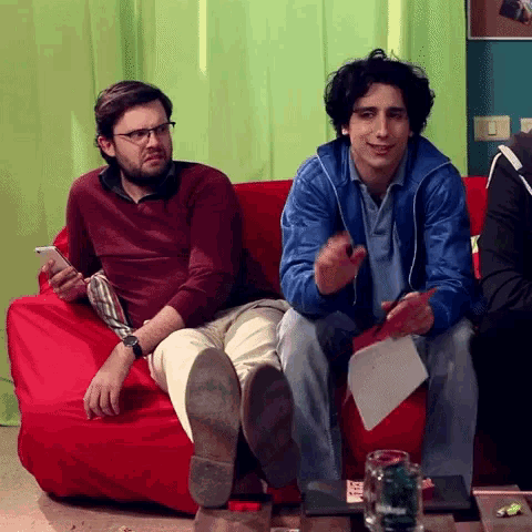 two men are sitting on a red couch and one is holding a piece of paper