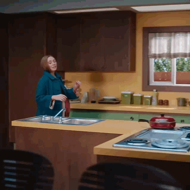 a woman in a blue coat is standing in a kitchen near a sink