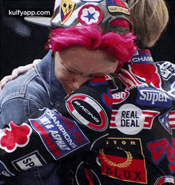 a man with pink hair is hugging another man wearing a jacket with patches on it .