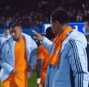 a man in a blue adidas jacket is pointing at another man in an orange shirt .