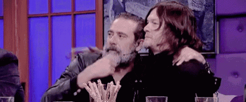 two men are sitting at a table with their arms around each other . one of the men has a beard .