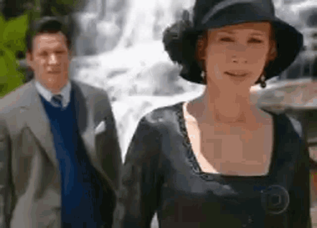 a man and a woman are standing in front of a waterfall . the woman is wearing a hat and a black dress .