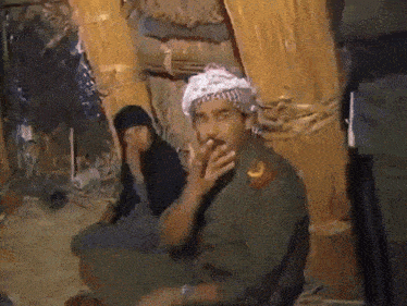 a man in a military uniform is smoking a cigarette while a woman sits behind him