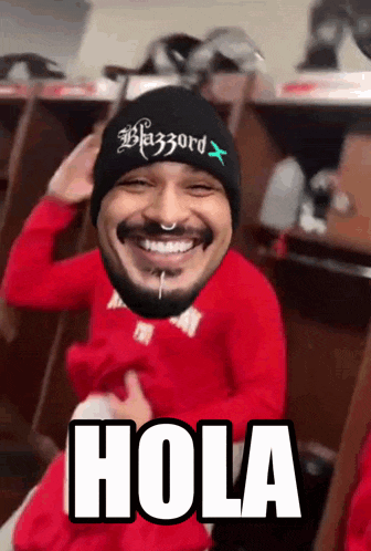 a man wearing a red shirt and a black beanie is smiling and says hola