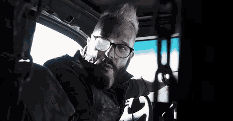 a man with glasses and a beard is sitting in the driver 's seat of a car .