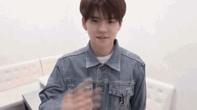 a young man wearing a denim jacket is sitting on a couch and giving a thumbs up .