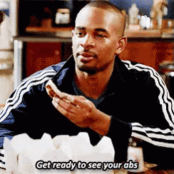 a man is sitting at a table eating a sandwich and says get ready to see your abs