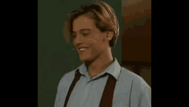 a young man wearing a blue shirt and red suspenders is smiling while standing in front of a green wall .