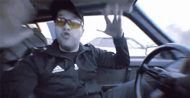 a man wearing sunglasses and a hat is driving a car and making a peace sign .