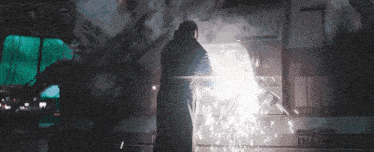 a man in a black coat is standing in front of a machine with a light coming out of it and sparks flying .