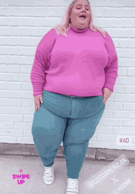a woman wearing a pink sweater and blue jeans is standing in front of a white brick wall