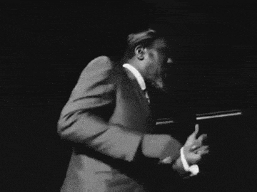 a black and white photo of a man in a suit and hat walking in the dark .