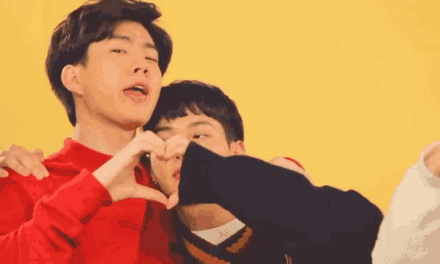 two young men are making a heart shape with their hands against a yellow background .