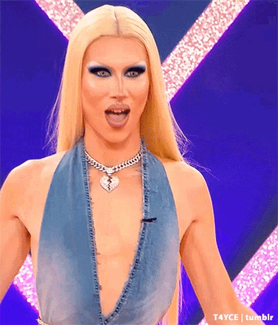 a drag queen with blonde hair and blue eye makeup is wearing a denim top and a heart necklace .