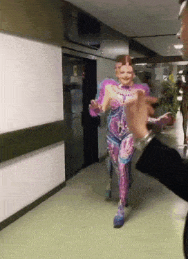 a woman in a pink and blue costume is walking down a hallway with a man pointing at her