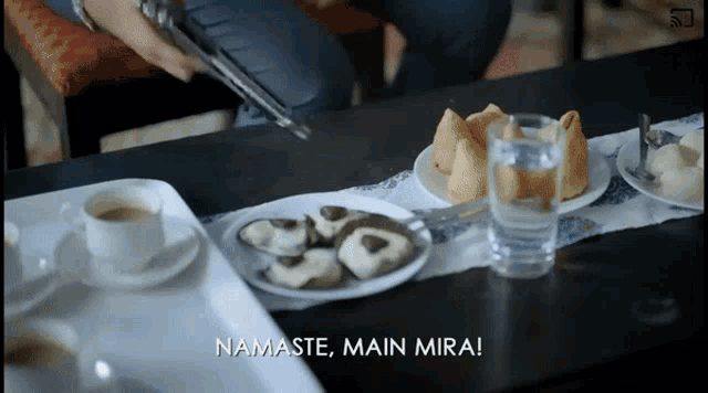a person is sitting at a table with plates of food and a cup of coffee and a glass of water and says namaste main mira