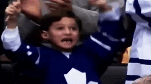 a young boy wearing a blue and white hockey jersey is sitting in the stands with his hands in the air .