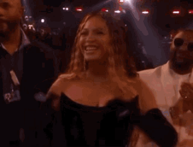 a woman in a black dress is smiling while standing next to a man in a white suit in a crowd .