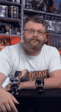 a man wearing a quake t-shirt sits in front of a shelf full of toys