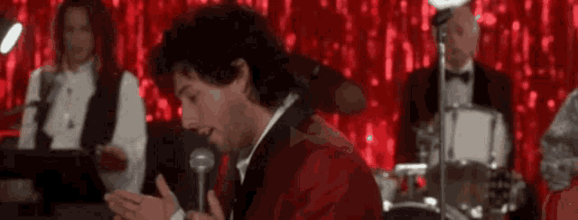 a man in a red tuxedo is singing into a microphone in front of a red curtain .